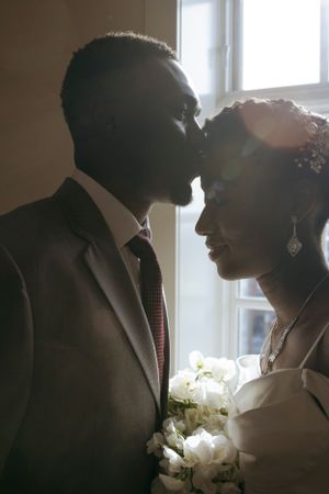 Silhouetted couple shares tender moment near window with bridal bouquet.