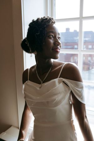 Elegant bride in white off-shoulder dress poses by window in natural light.