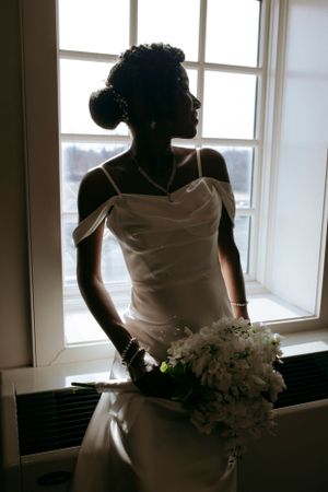 Silhouette of bride holding bouquet poses gracefully by window.