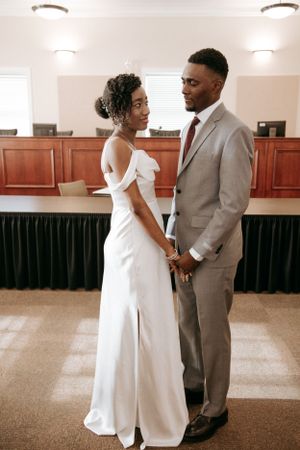 Couple in wedding attire holds hands while standing in formal reception room.