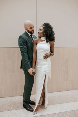 Elegant couple poses in formal wedding attire against a neutral wall in courthouse setting.