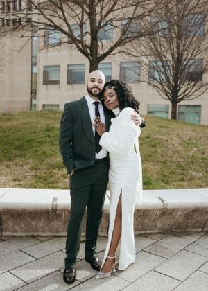 A couple in formal attire poses outdoors, with her wearing a white dress and him in a dark suit.