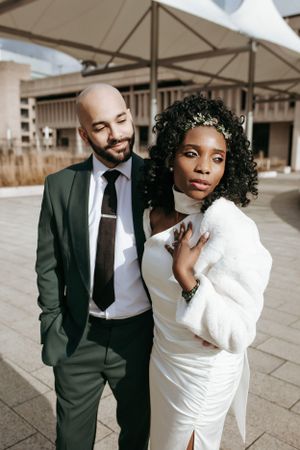 A stylish couple poses outdoors, with her in a white dress and him in a dark suit.
