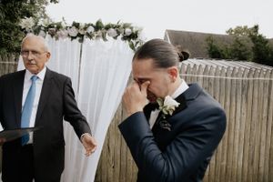 Emotional groom wiping tears during outdoor wedding ceremony while officiant stands nearby.