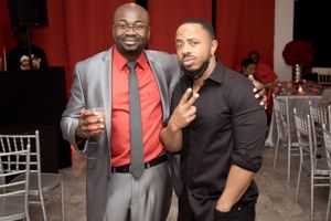 Two men in formal attire posing together at an event with silver chairs in the background.