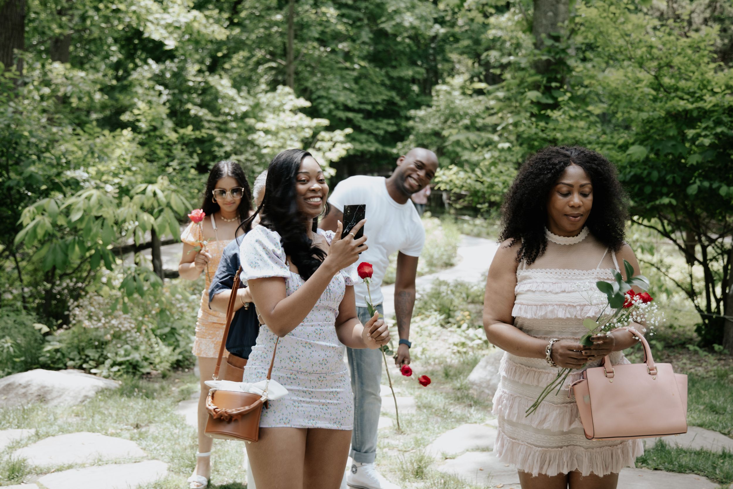 Group walking through wooded path carrying bags and flowers.
