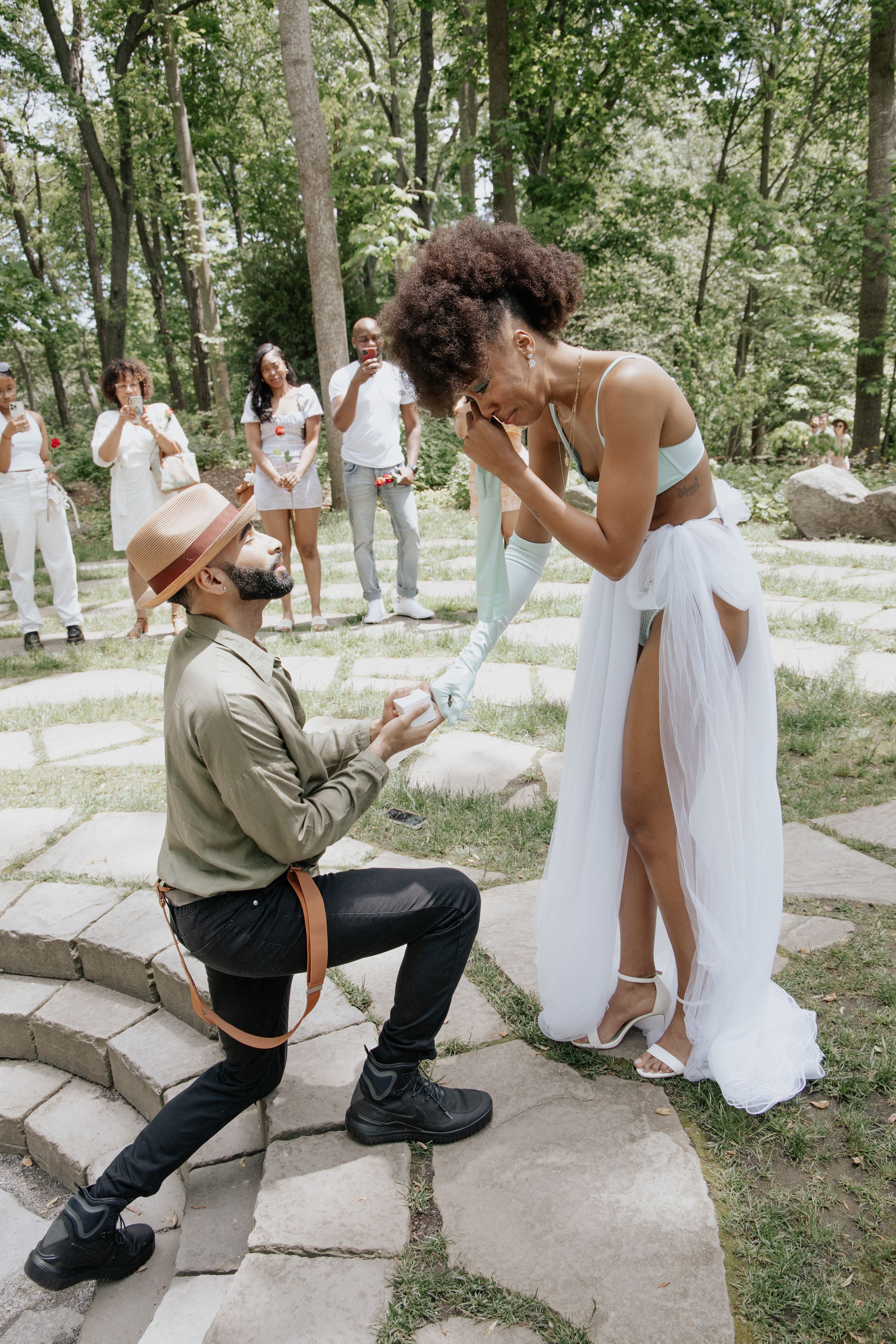 A romantic outdoor proposal scene on stone steps surrounded by trees and onlookers wearing all white.