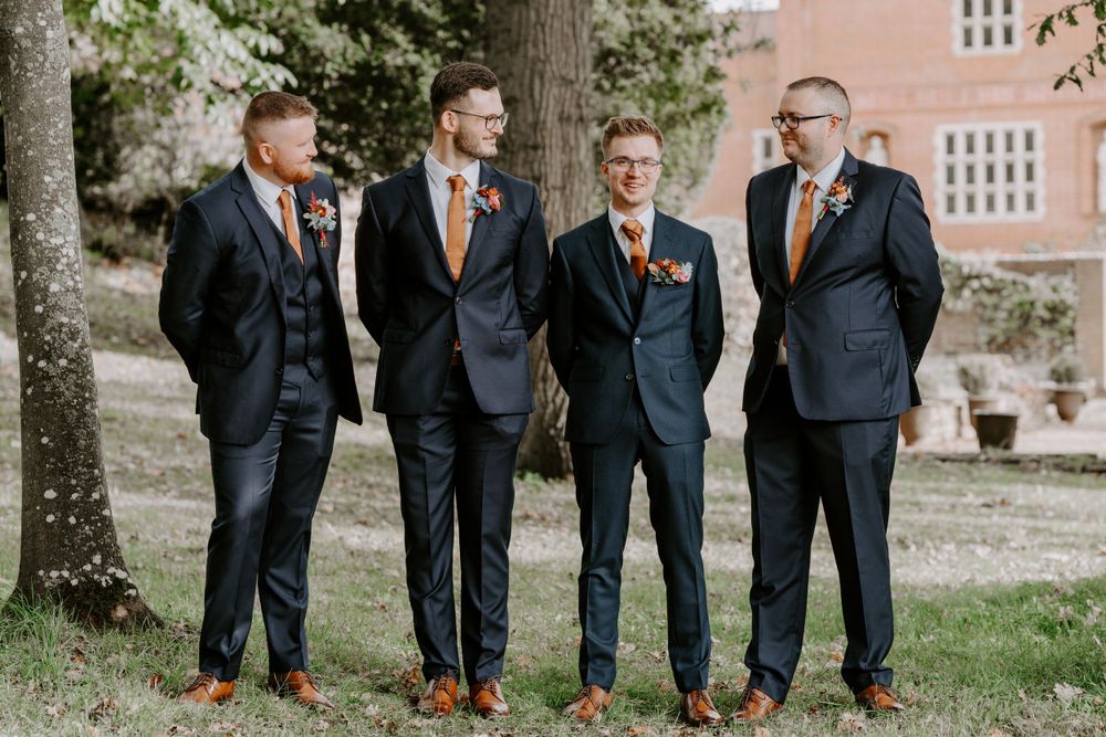 A groom and his 3 groomsmen are stood in the grounds of oxnead hall in Norfolk. They are looking at each other and smiling.