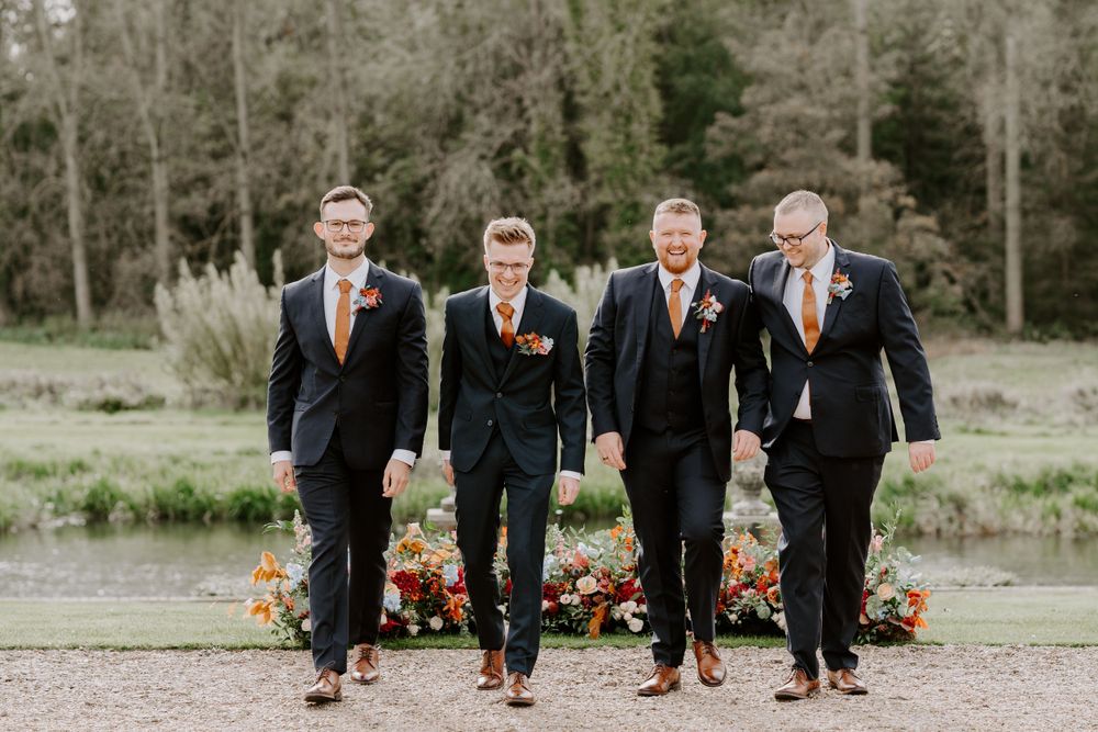 A groom and his 3 groomsmen are walking towards the camera. They are looking at each other and smiling.