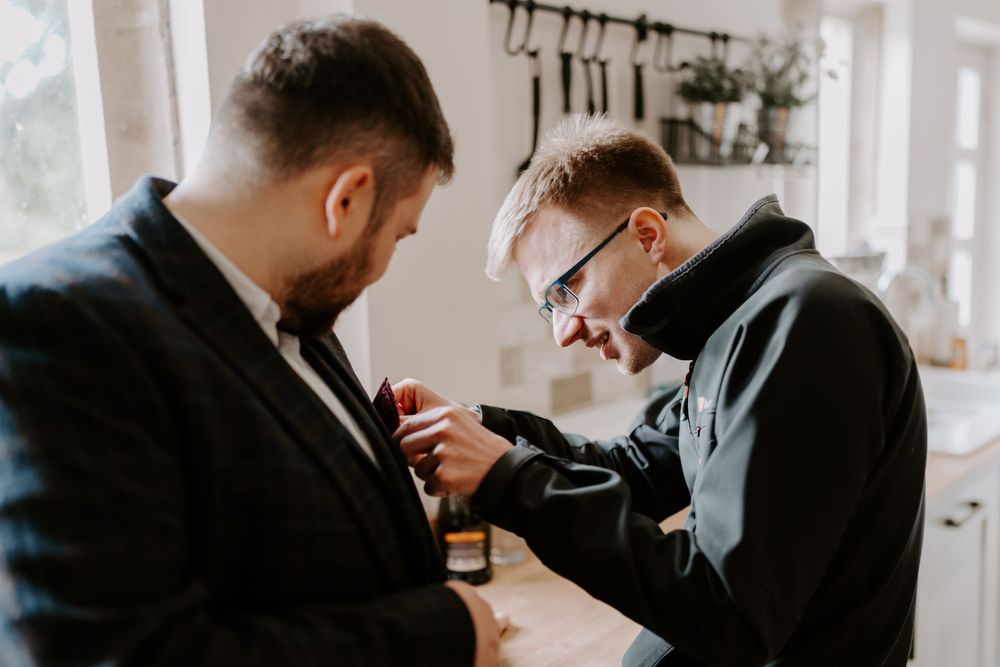 Groom is pinning a button hole onto his groomsman’s suit lapel.