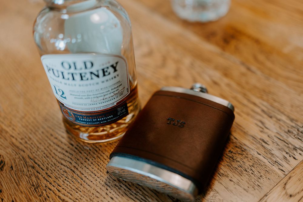 A close up of a brown leather and chrome hip flask beside a bottle of whiskey.