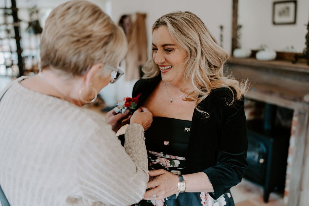 The mother of the groom is having her button hole pinned on by her mum. She is looking at her mum and smiling.