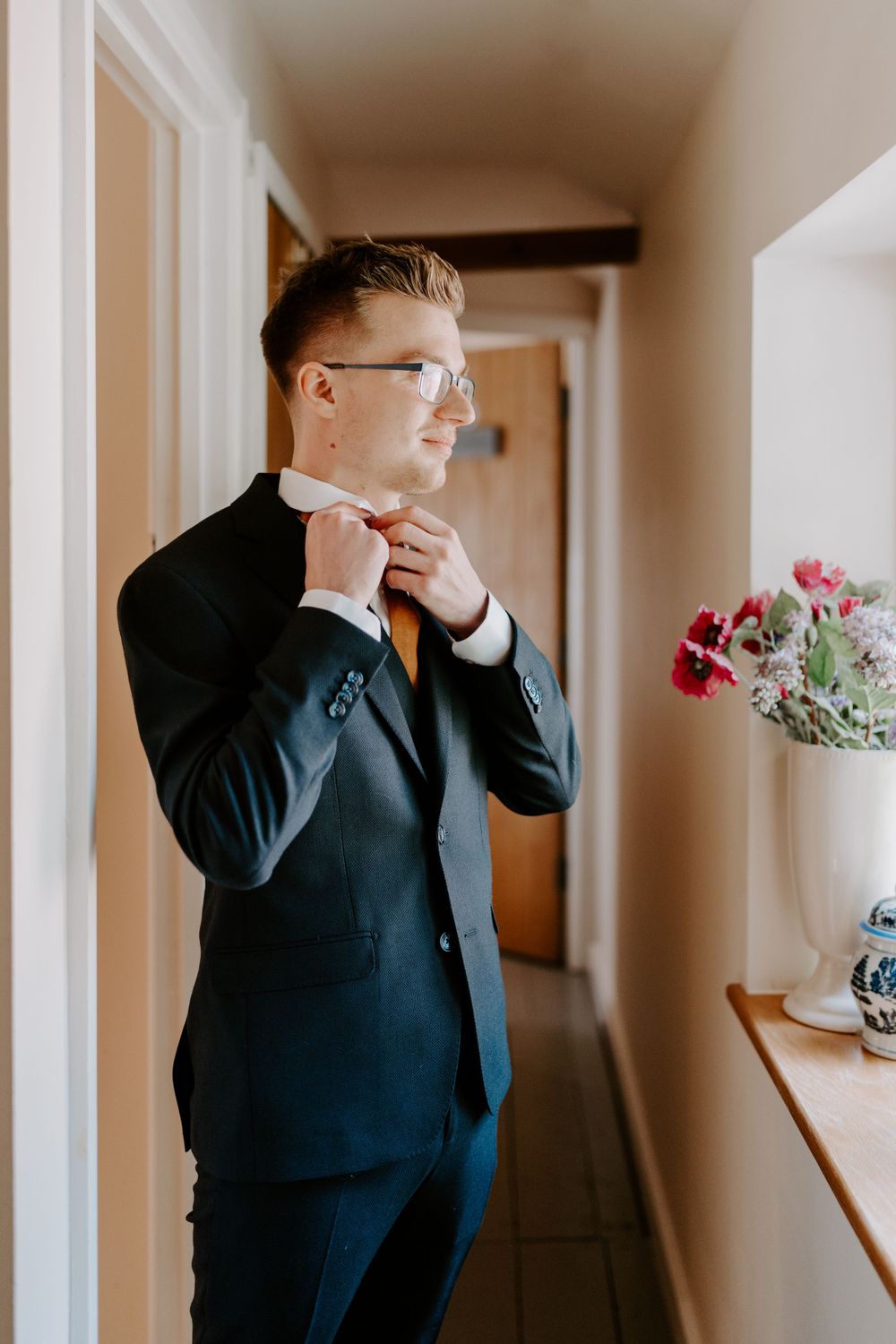 Groom is standing in front of a window and is looking out of the window. He is tying he’s tie which is burnt orange and wearing a navy suit.