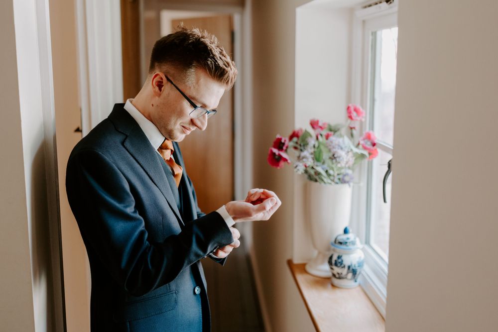 Groom is stood in front of a window and is adjusting his cufflinks, looking down at them. He is wearing a navy suit and a burnt orange tie.