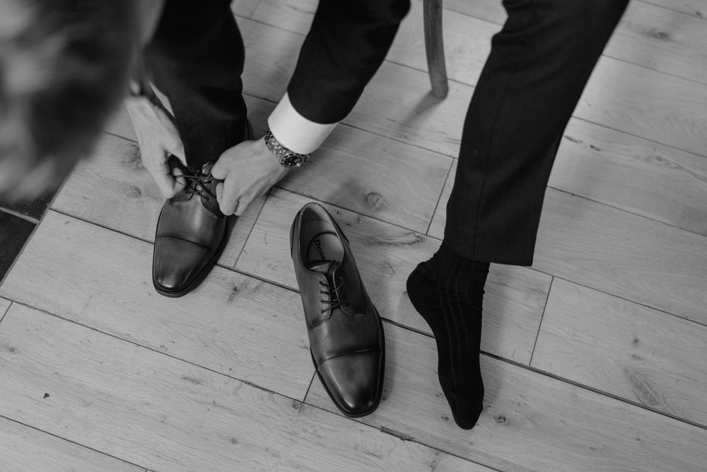 A black and white photo, a close up of a groom putting his shoes on and tying his laces. Just his hands and feet are in shot.