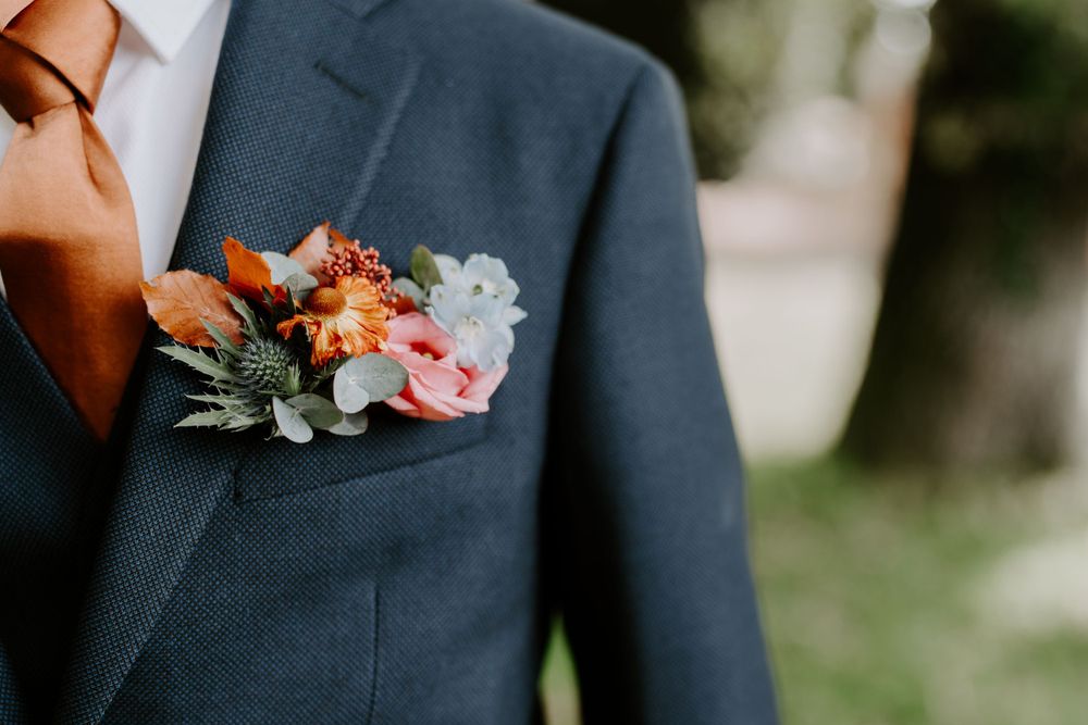 A close up of a bottom hole. Flowers are pink and orange with greenery. The grooms suit is navy.