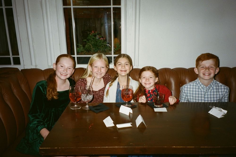 Children sitting at a wooden table with paper crafts during what appears to be a holiday gathering.