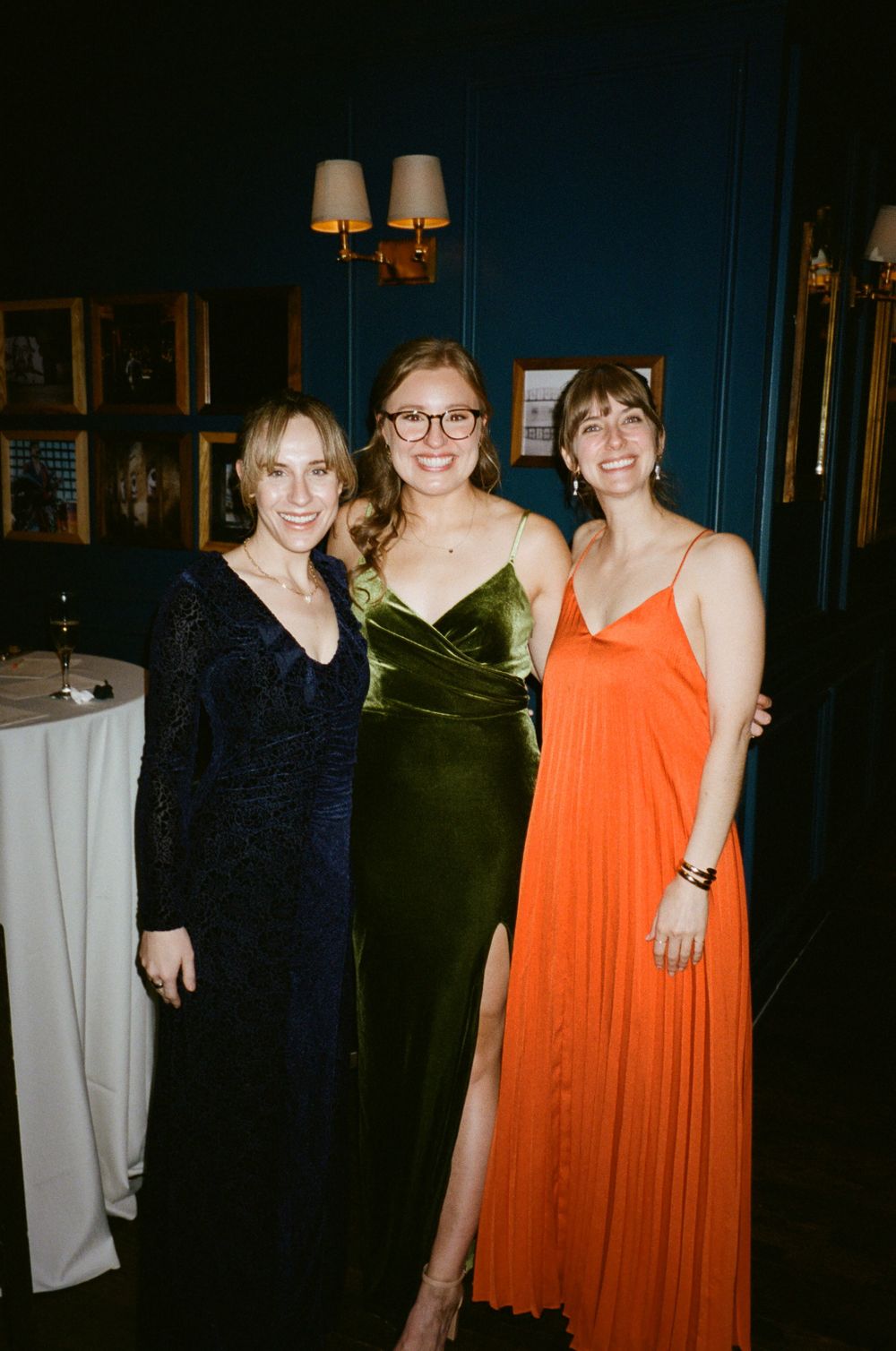 Three women in formal dresses pose together at an elegant event venue.