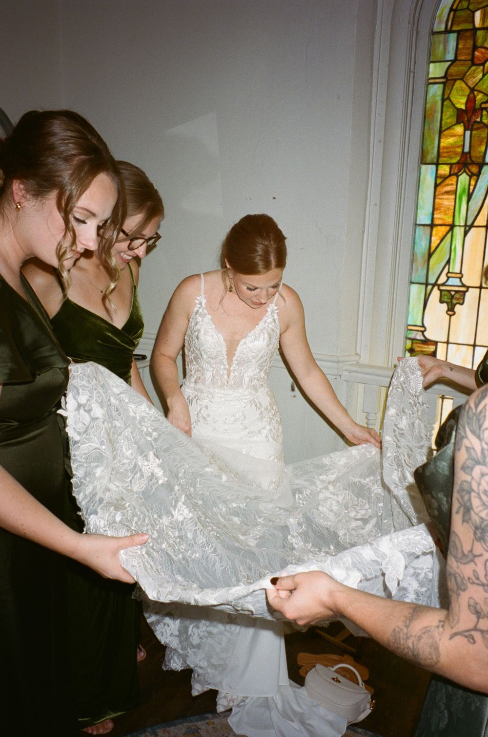 Bridal party helping with wedding dress adjustments near stained glass window.