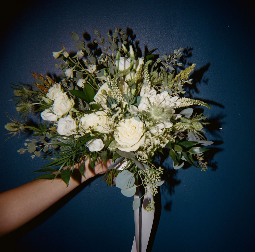 A lush white and green wedding bouquet with roses and mixed flowers held against a blue background.