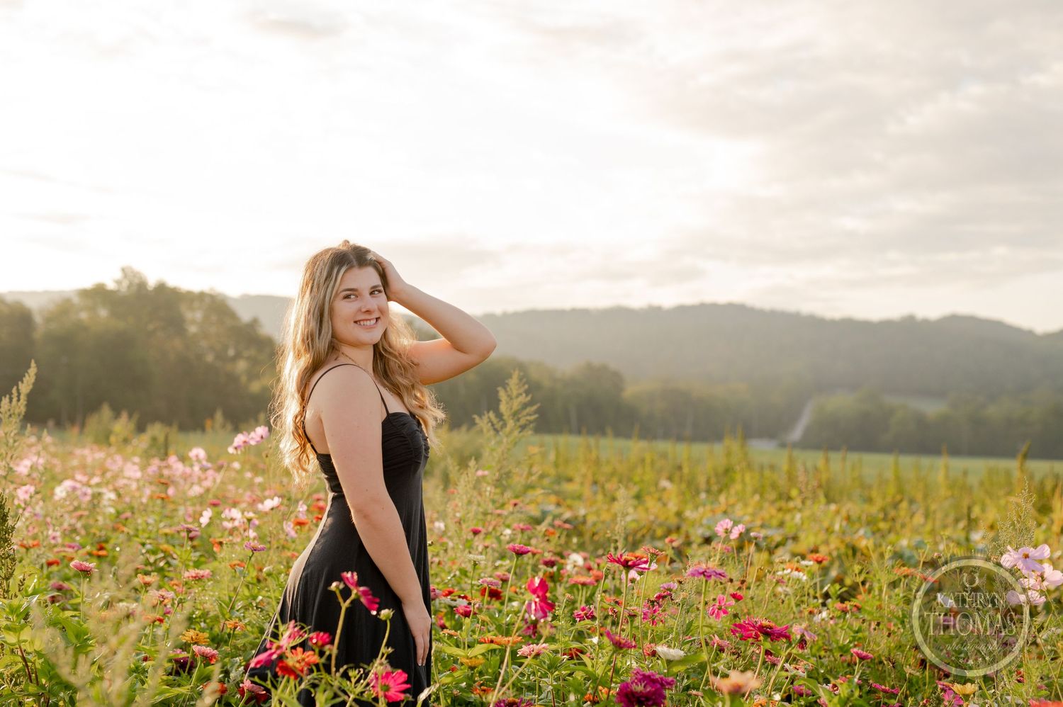 A person in a black dress poses in a field of pink wildflowers at sunset.