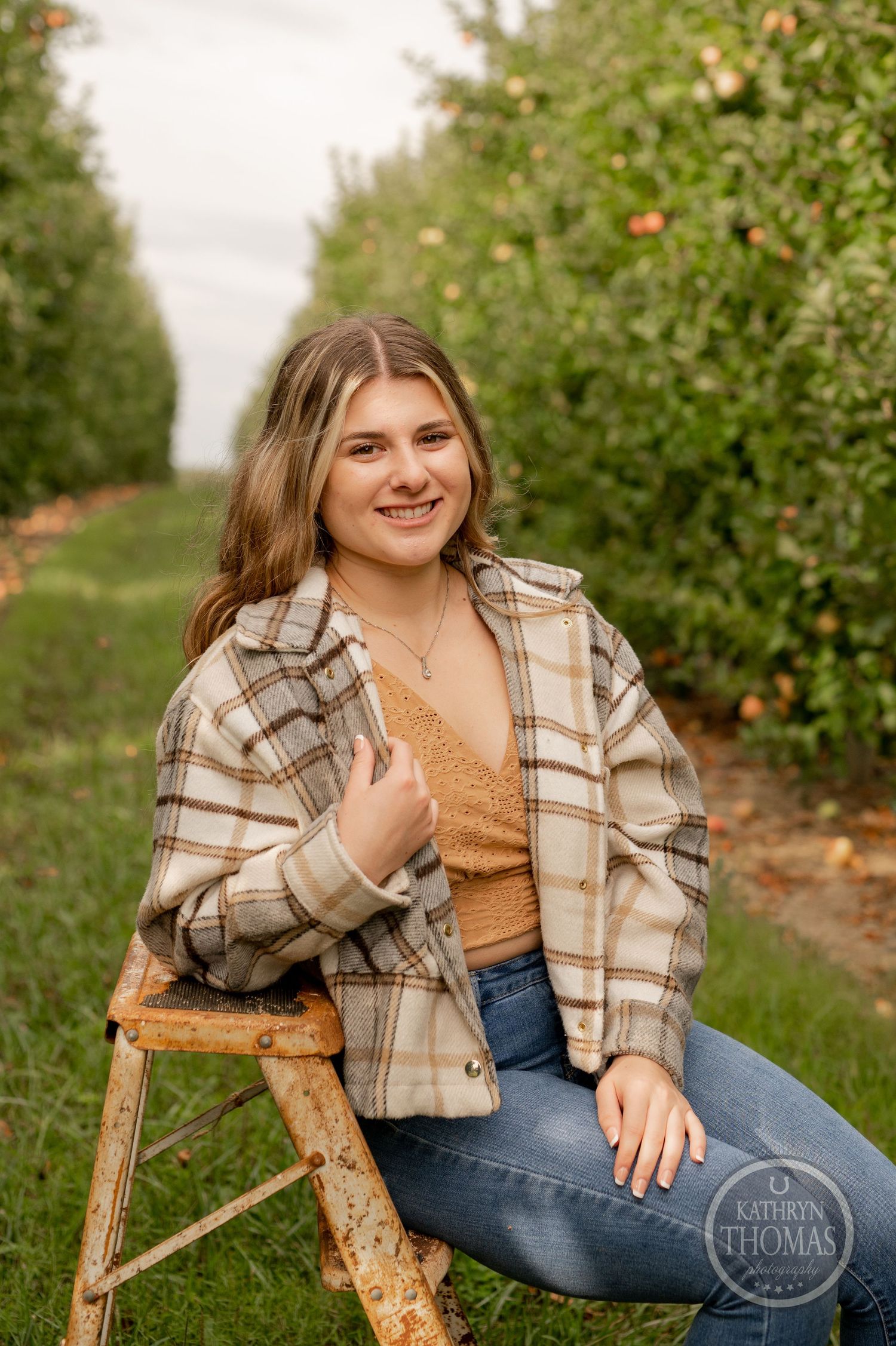 Person in plaid jacket sits on rustic stool between rows of apple trees in orchard.