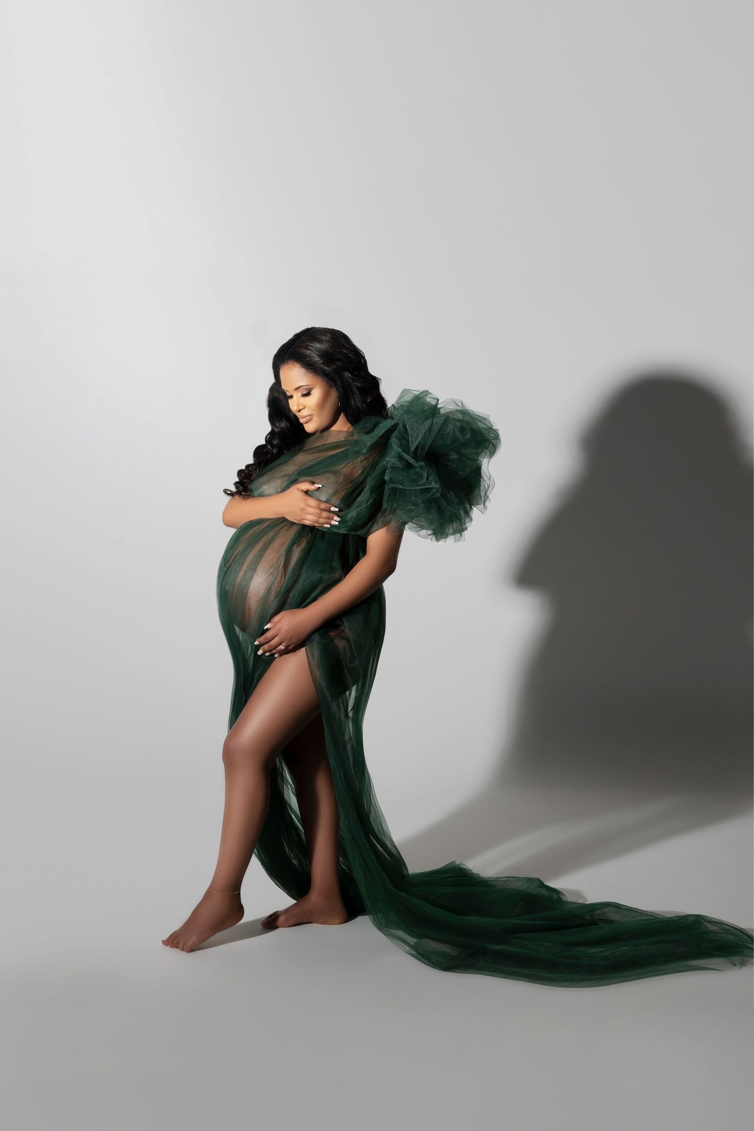 Elegant maternity photograph with flowing emerald green tulle gown against light gray studio backdrop with dramatic shadow.
