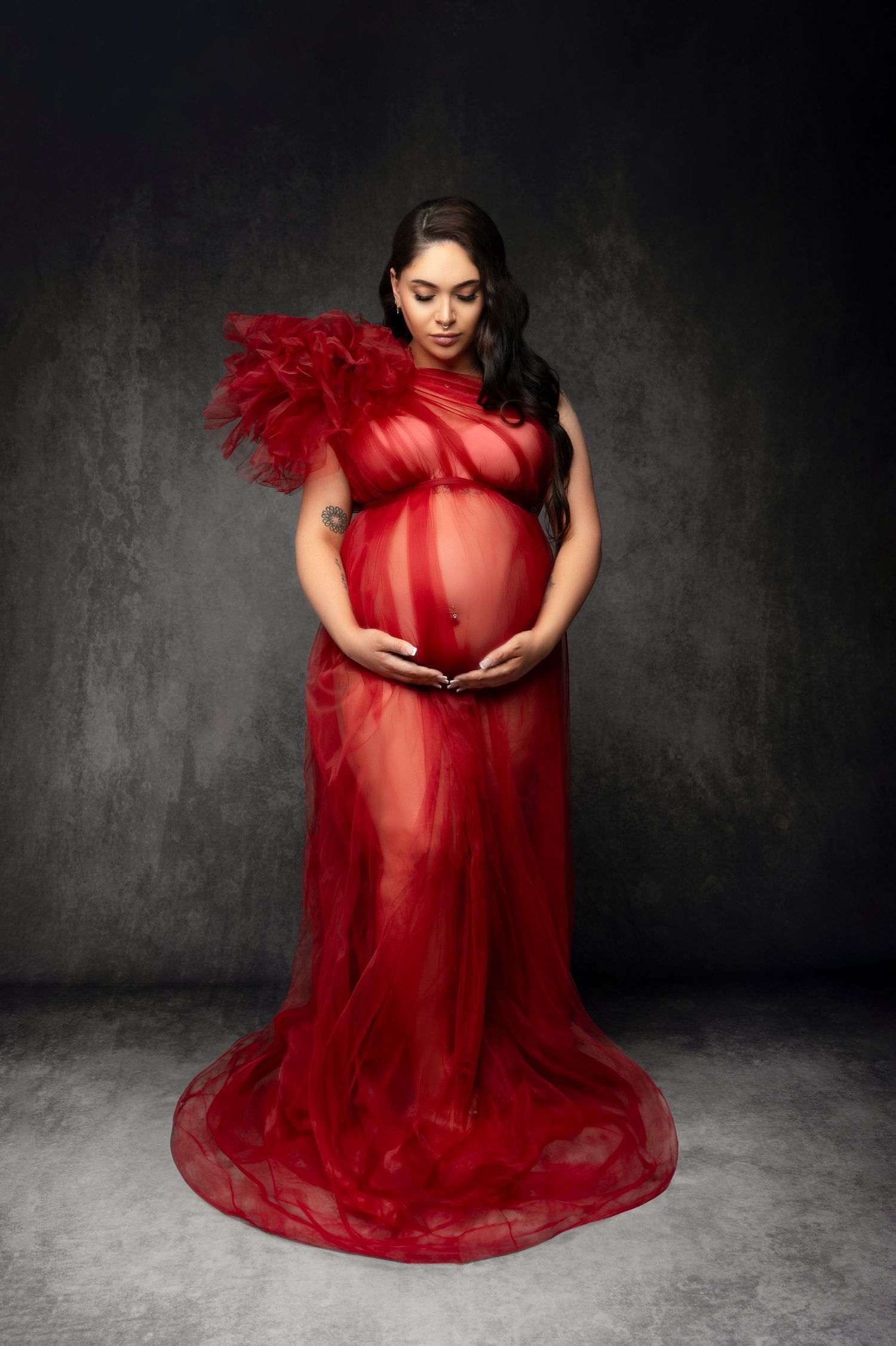 Maternity photo featuring an elegant red sheer gown with feather details against a dark dramatic background.