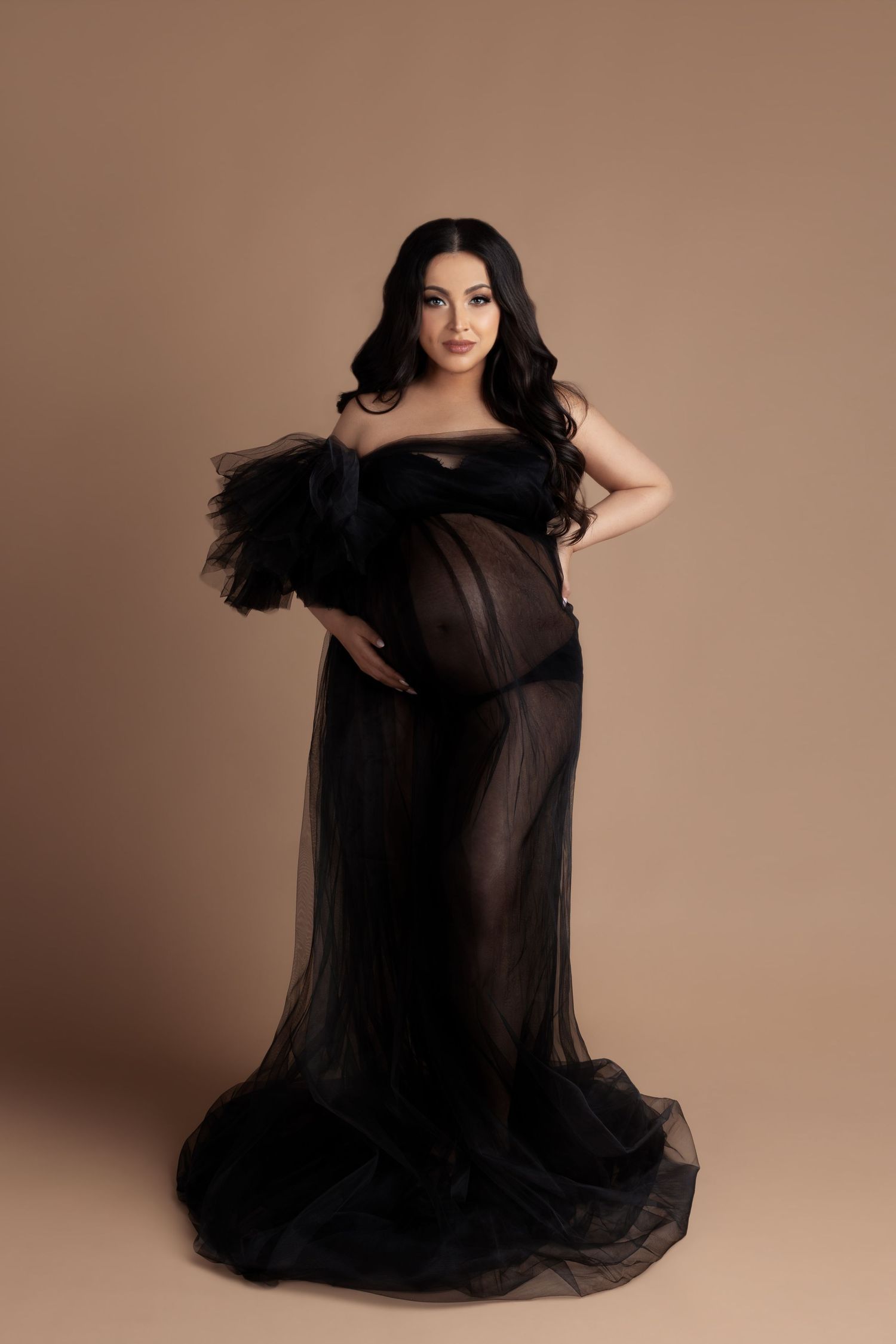 Elegant maternity portrait featuring a flowing black tulle gown against a neutral beige backdrop.