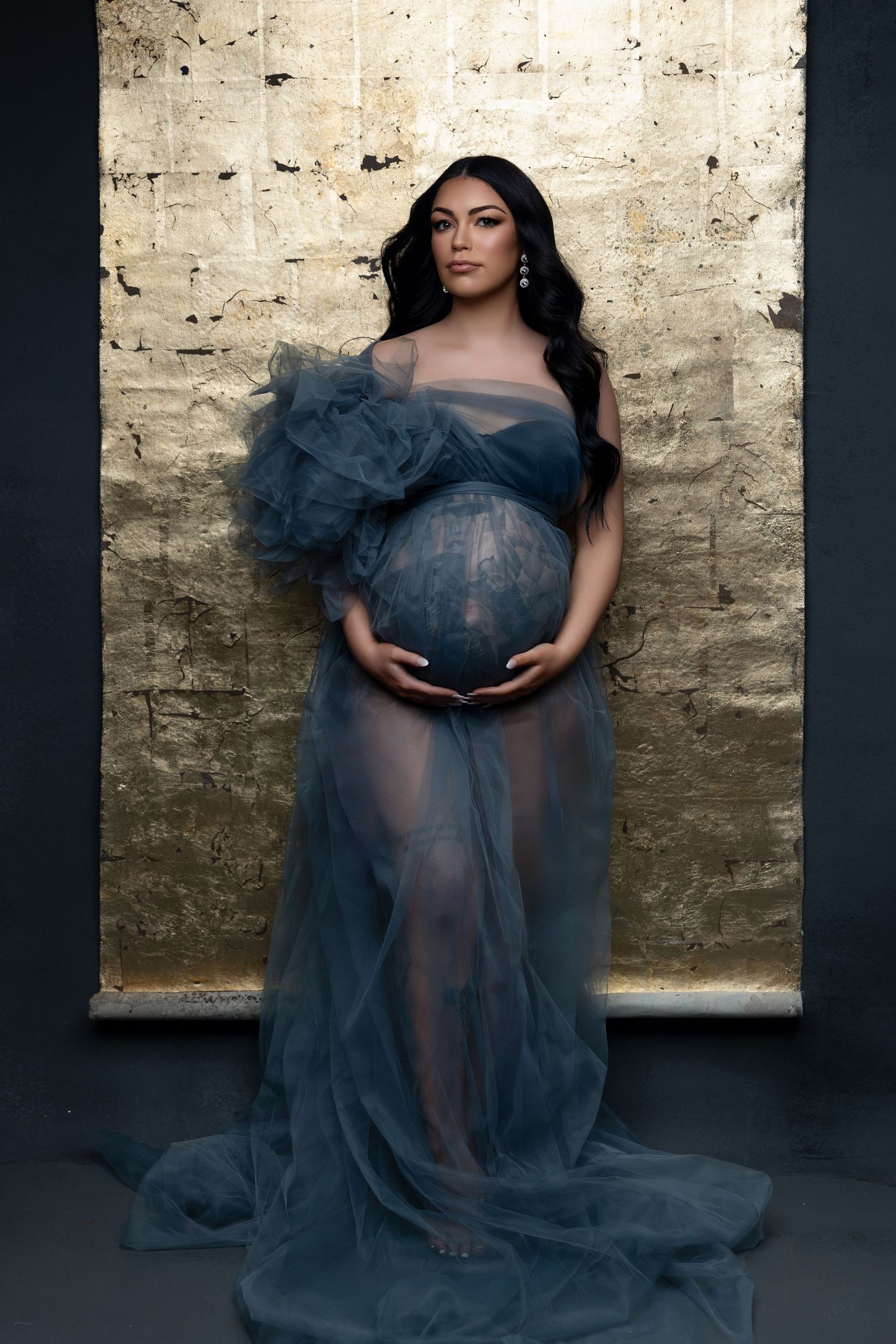 A figure poses in a flowing blue tulle maternity gown against a metallic gold background.