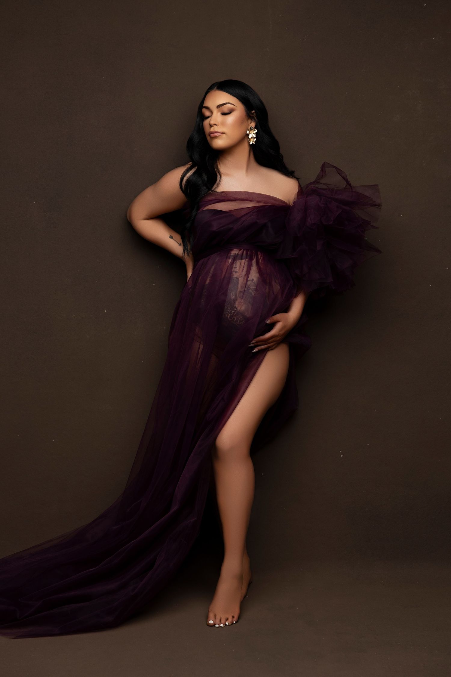Maternity photo featuring a flowing deep purple tulle gown against a dark brown backdrop in elegant studio lighting.