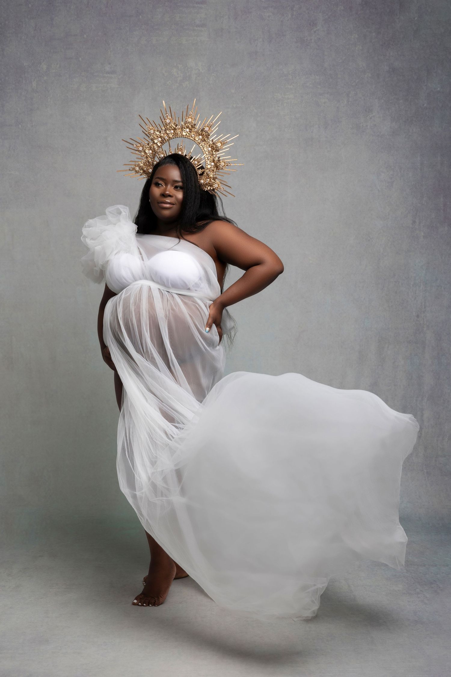 An elegant maternity portrait featuring flowing white fabric, a golden sun-inspired crown, and ethereal movement.