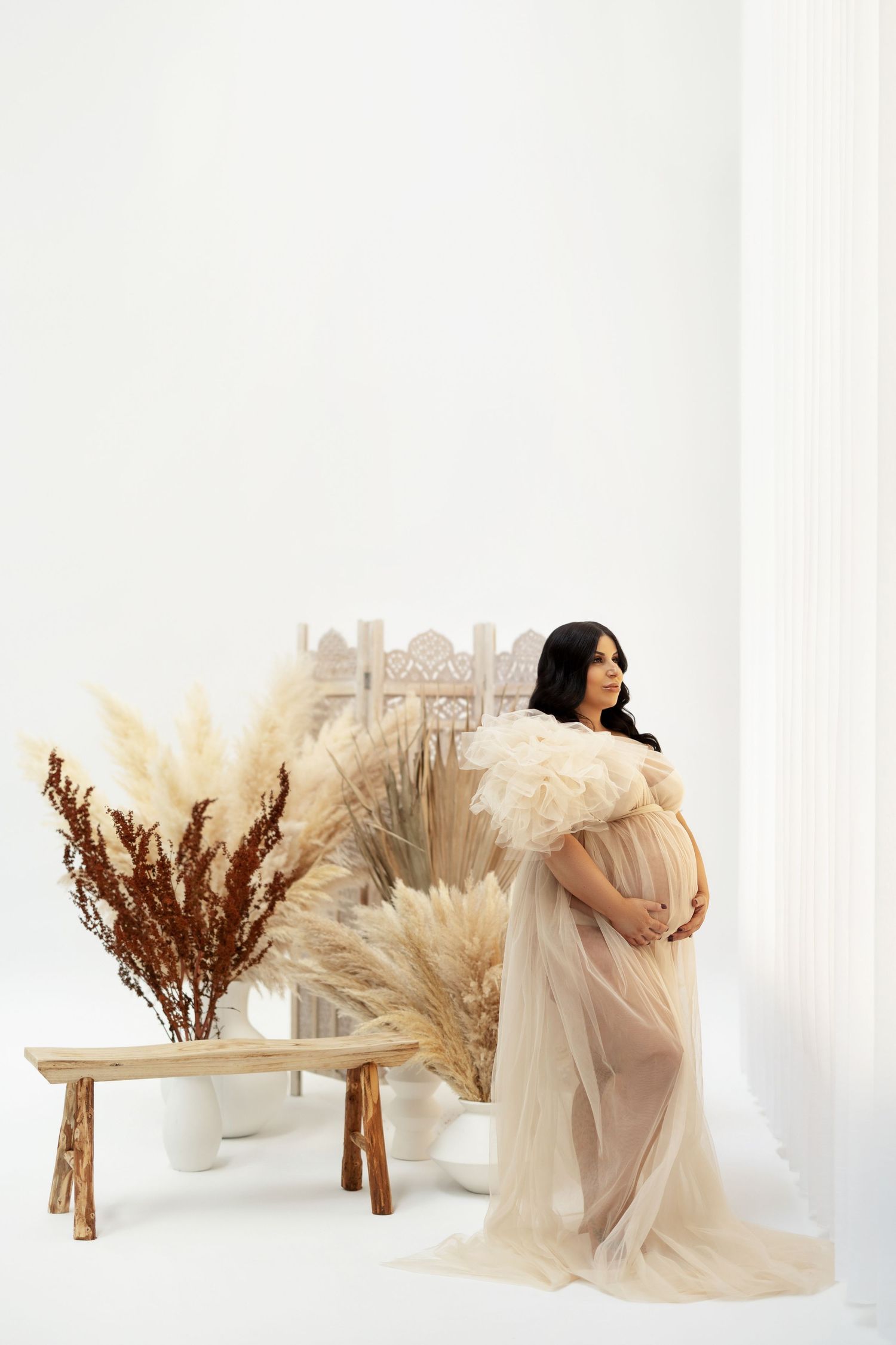 A maternity photoshoot featuring pampas grass and dried floral decorations against a white backdrop.