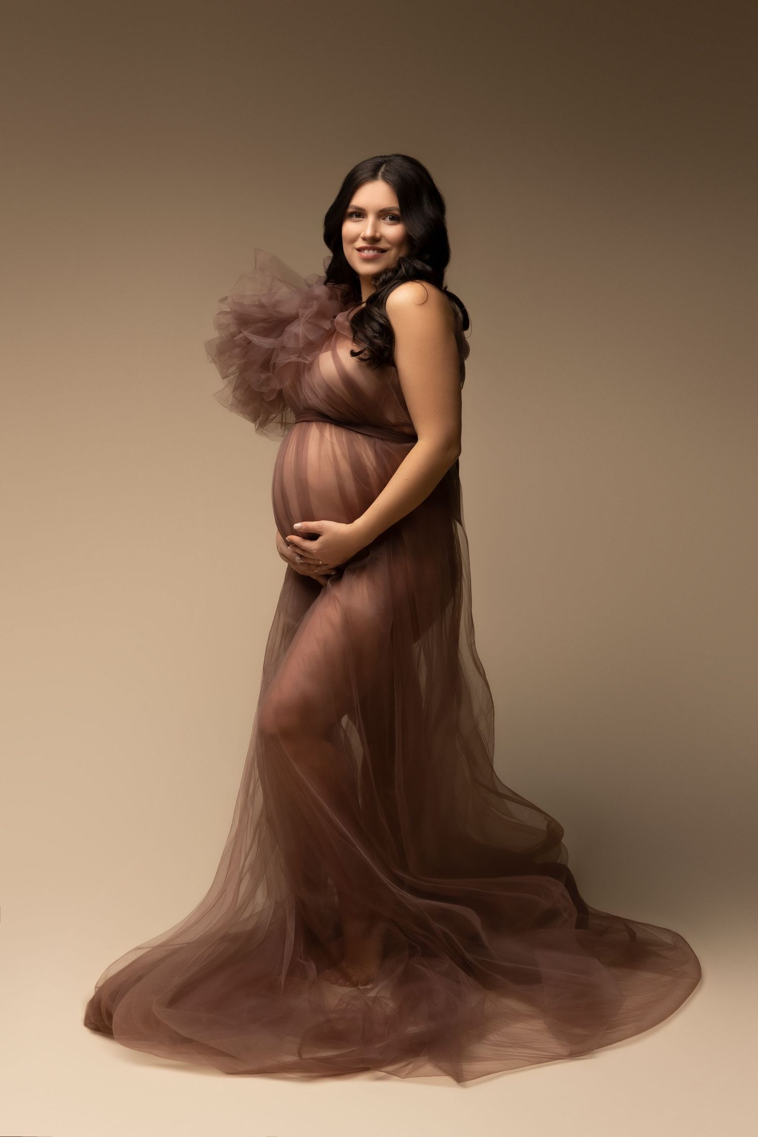 Maternity portrait in flowing brown tulle gown against neutral backdrop, highlighting an elegant pregnancy silhouette.