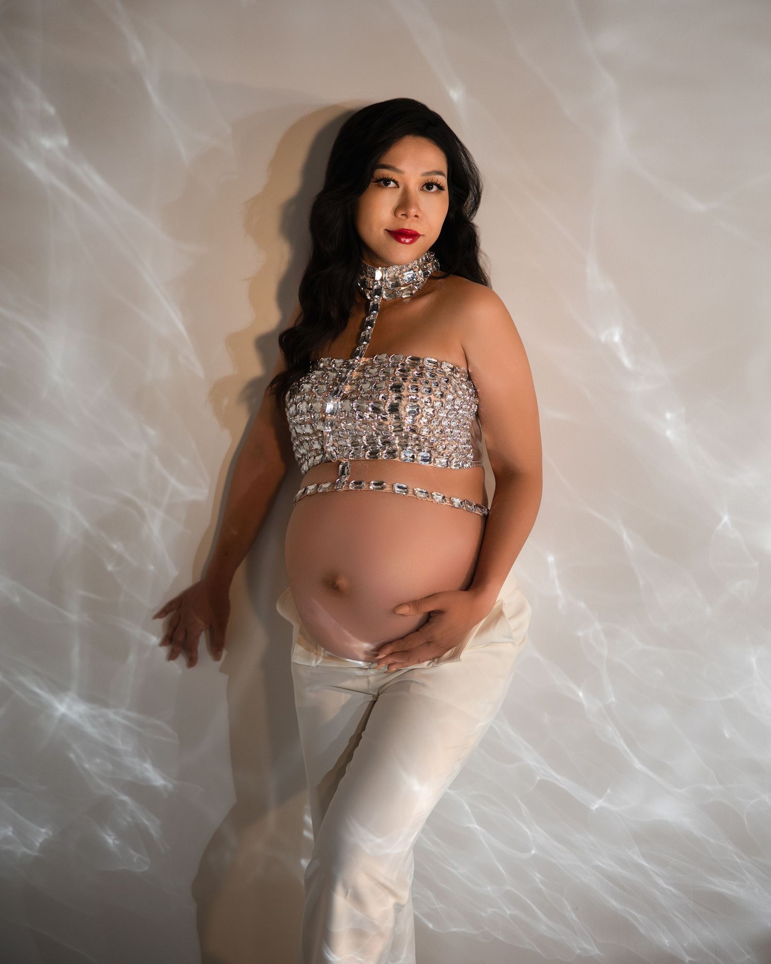 A pregnant woman in a sparkly silver crop top and white pants poses against a softly lit studio backdrop.