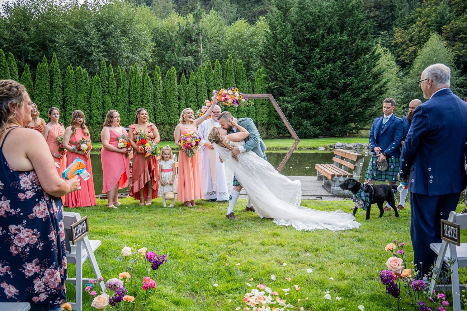 Outdoor garden wedding ceremony with guests seated on white chairs surrounded by evergreen trees.