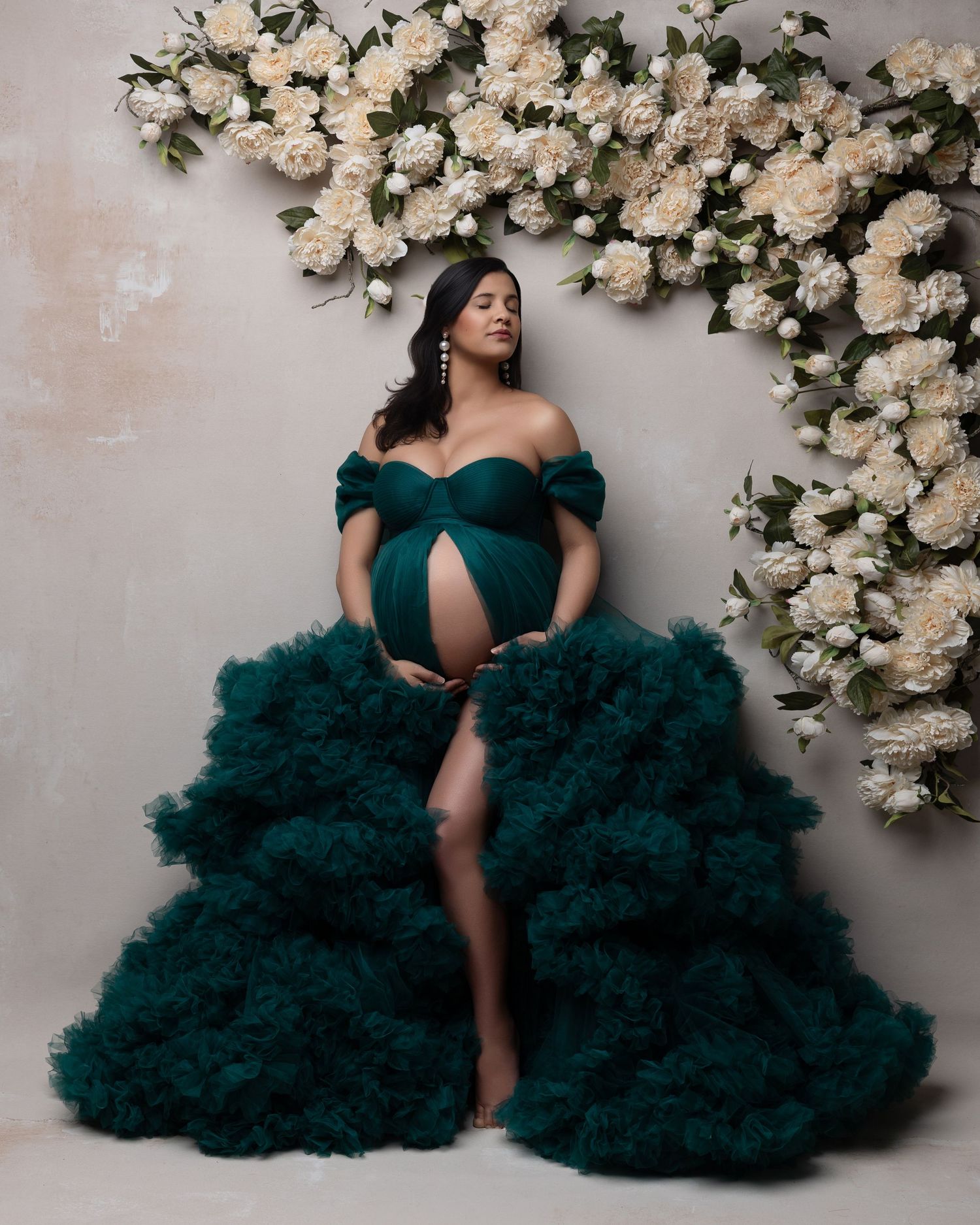 A woman in an elegant dark teal tulle gown poses against a wall adorned with white roses in a glamorous photoshoot.
