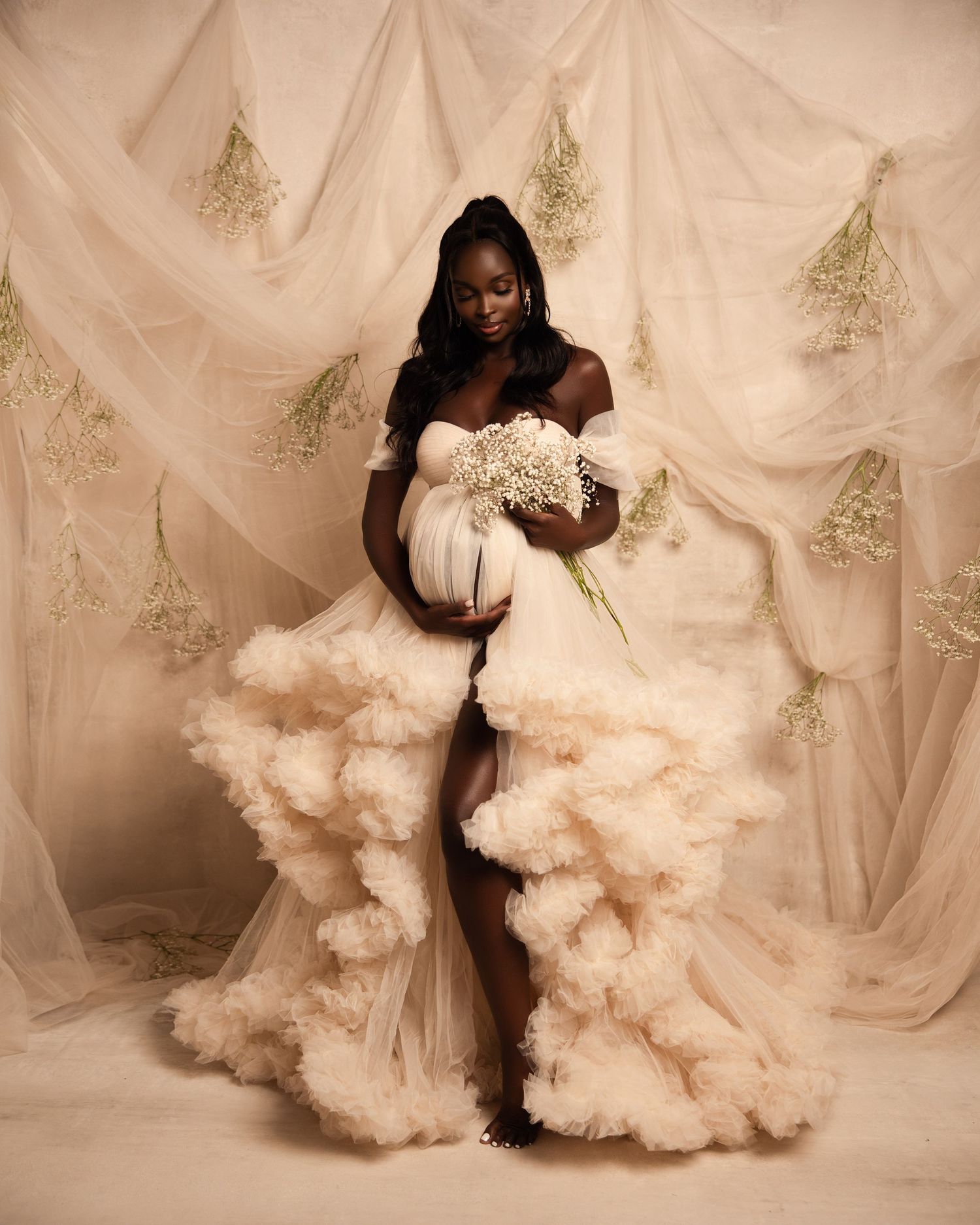 A maternity photoshoot featuring a dramatic ruffled white gown against a champagne-colored backdrop with delicate floral accents.