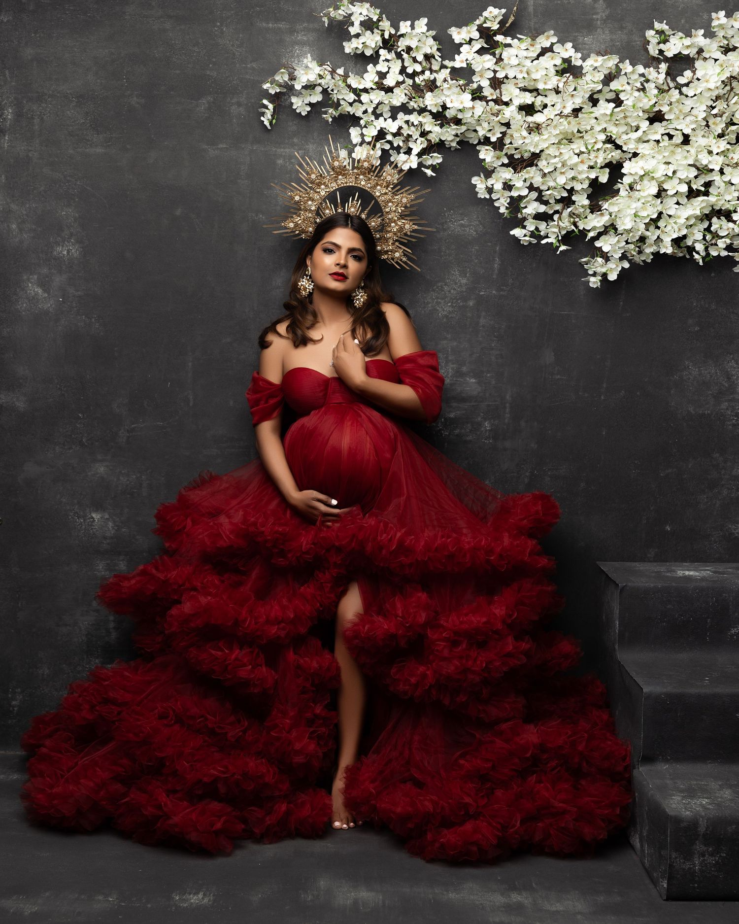 Dramatic fashion portrait with flowing deep red tulle gown against dark backdrop with white floral accents above.