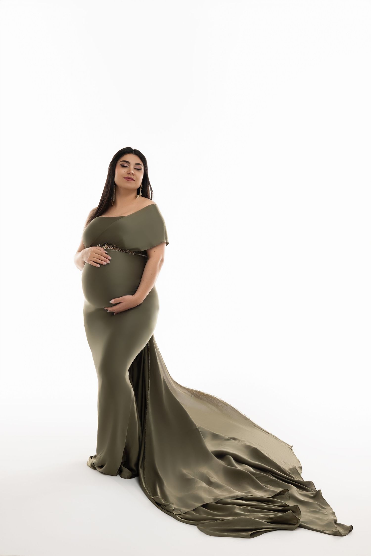 Elegant maternity photo featuring an olive-green flowing gown with off-shoulder design and dramatic train on white backdrop.