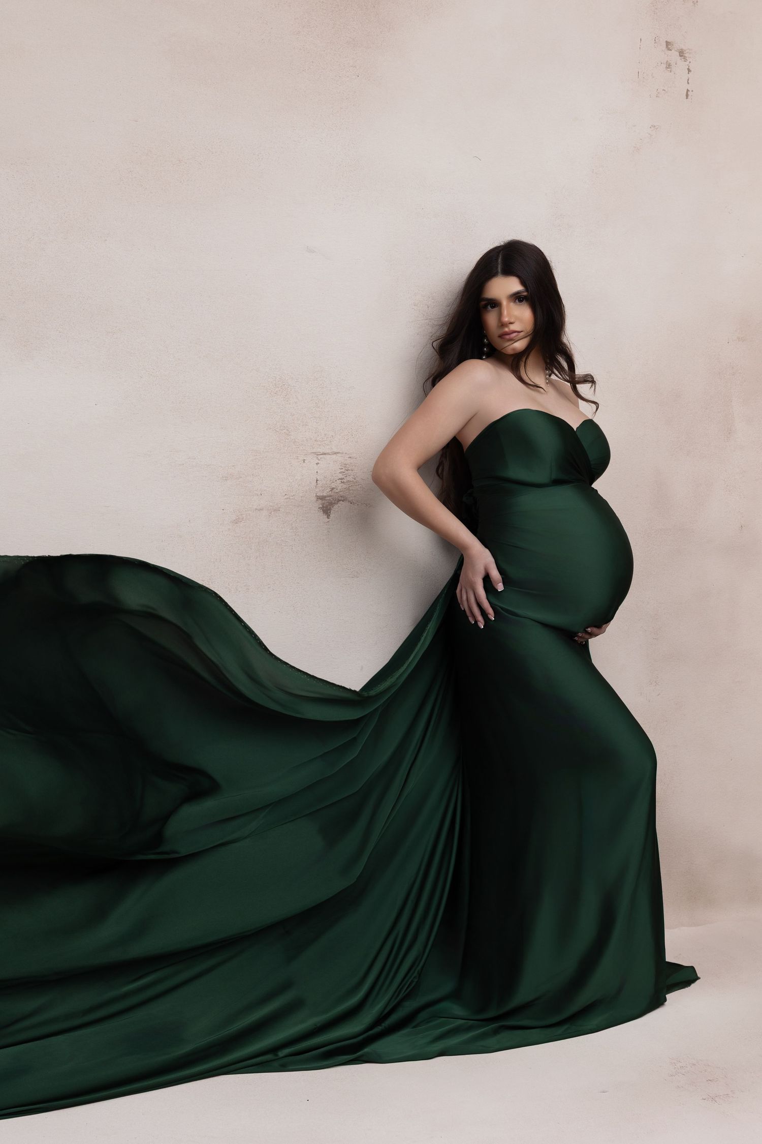 Pregnant woman in elegant emerald green silk gown with flowing train, posing against neutral background.
