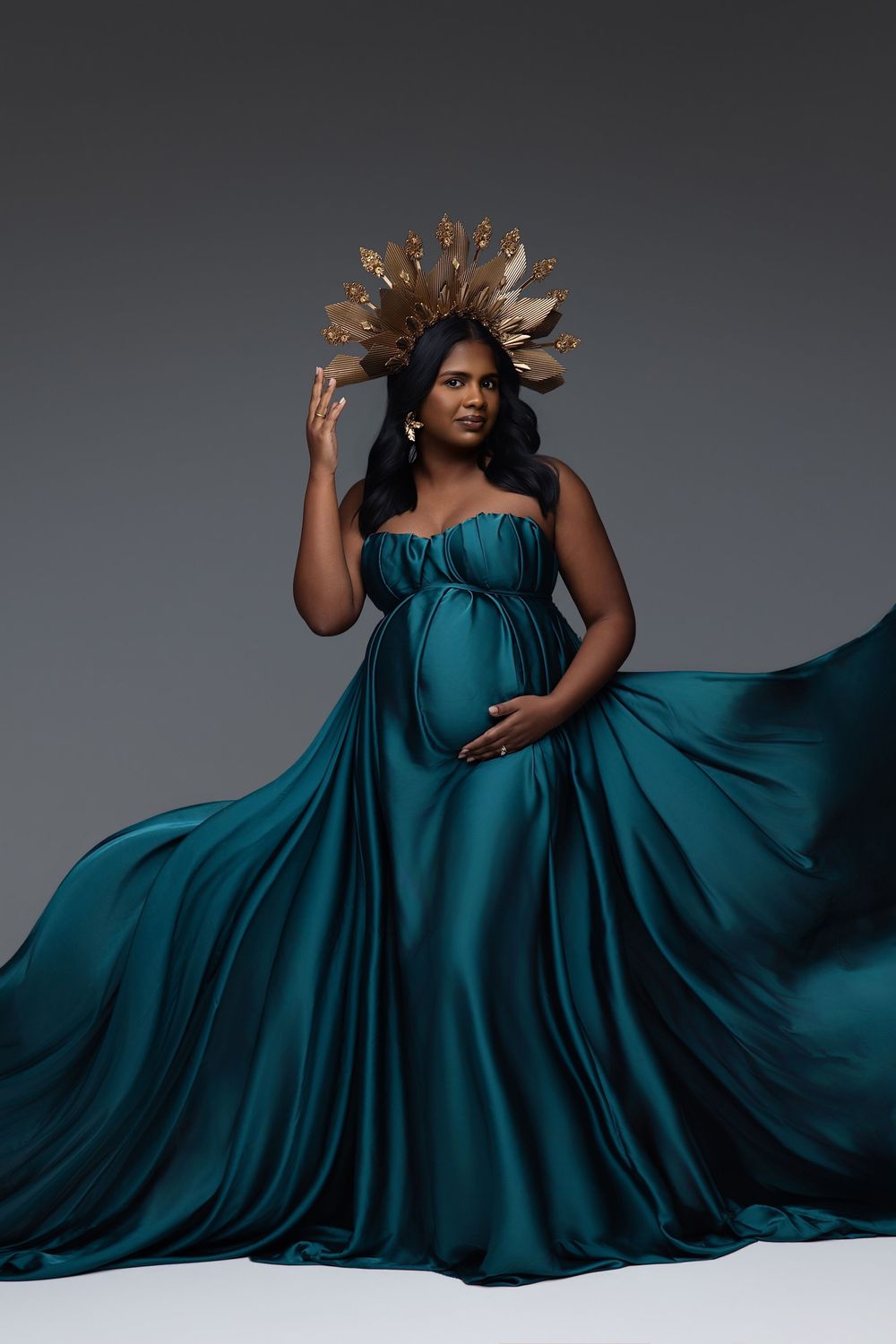 Pregnant woman in teal satin gown with gold crown headpiece poses gracefully against gray studio background.
