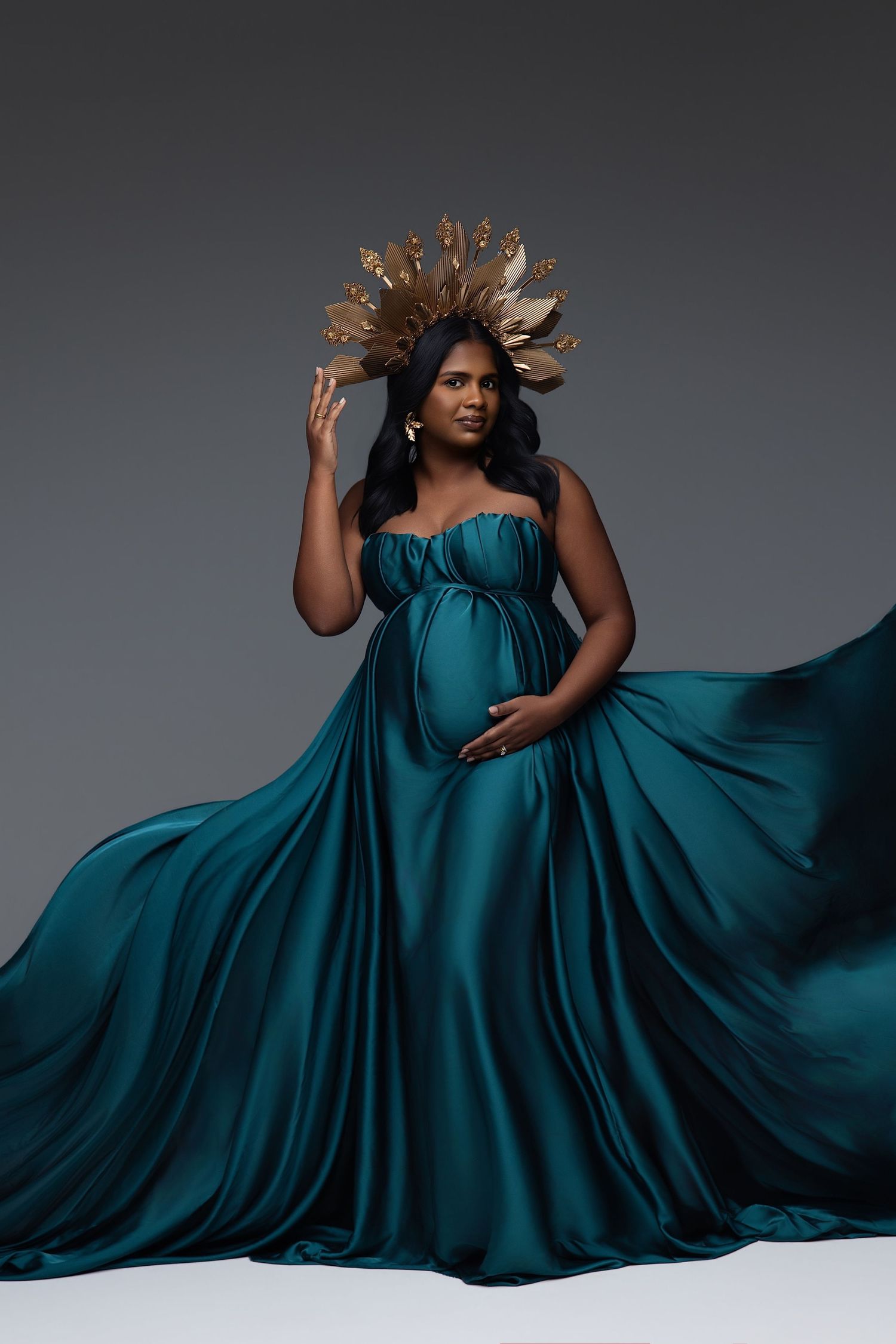 Pregnant woman in teal satin gown with gold crown headpiece poses gracefully against gray studio background.