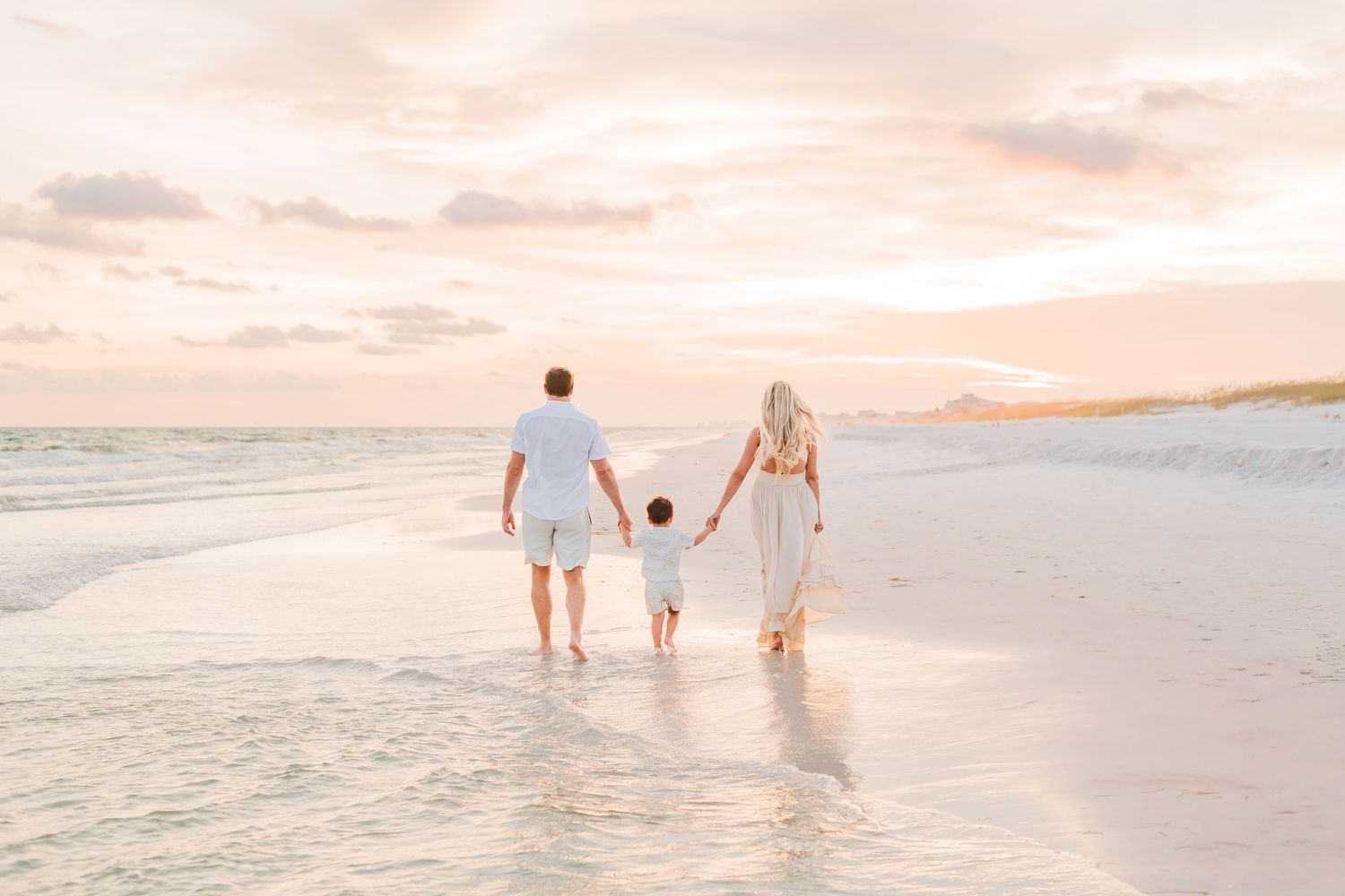 A couple walking together barefoot on a white sandy beach at sunset with waves crashing against the shore.