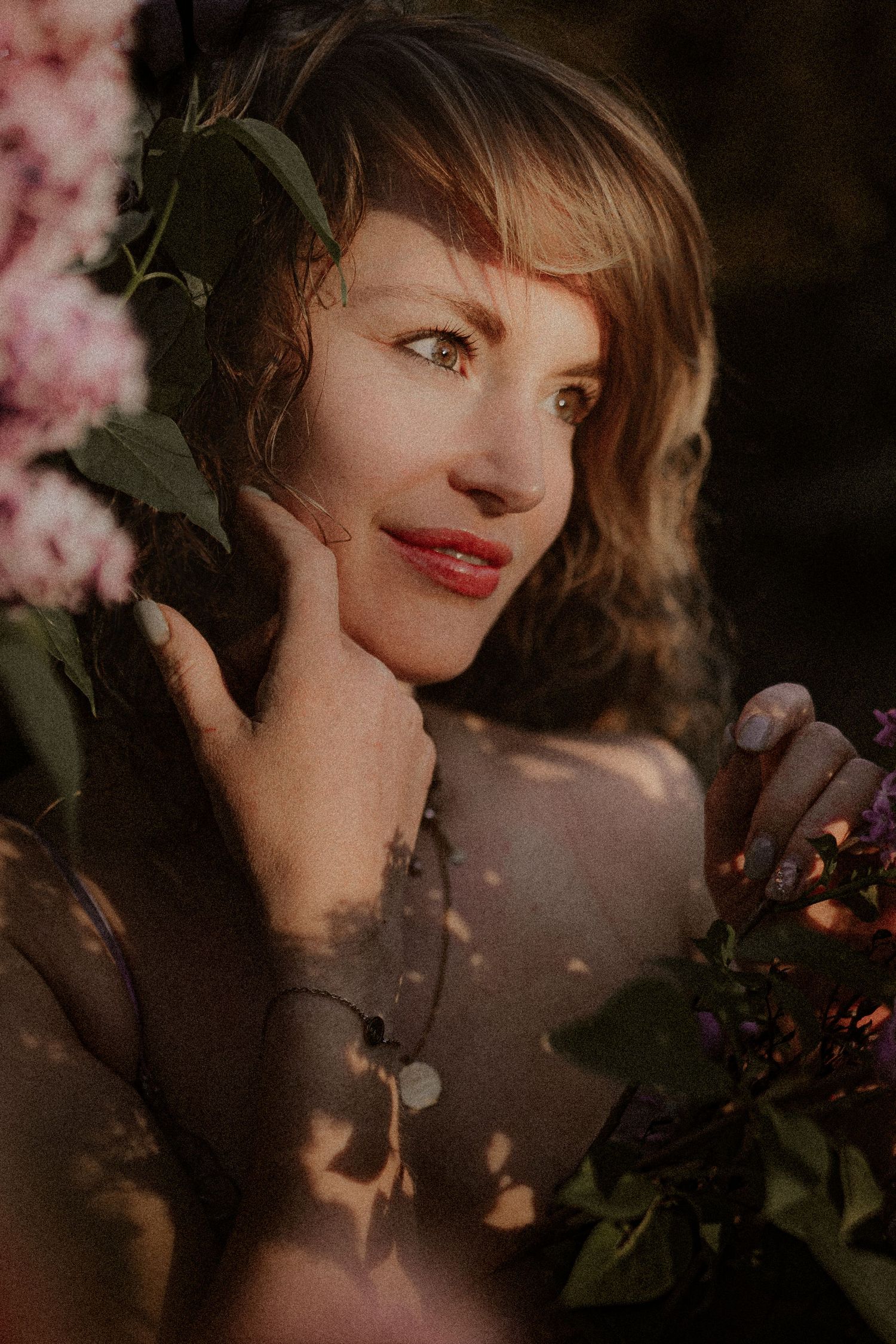 A woman with short hair and red lips poses gracefully among pink flowers and green leaves in warm golden light.