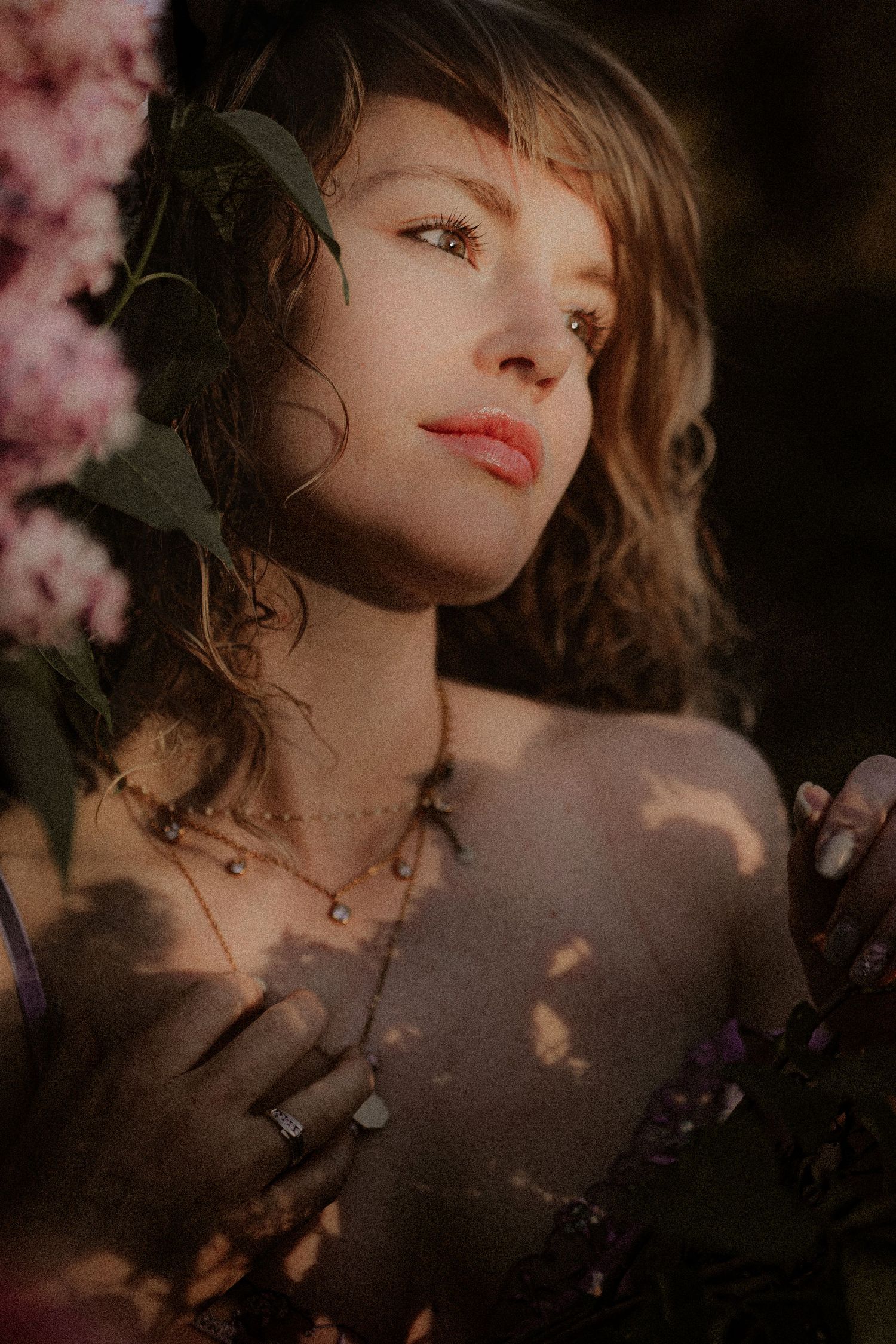 A woman with wavy hair poses among blooming pink and purple flowers, dappled sunlight casting soft shadows on her skin.