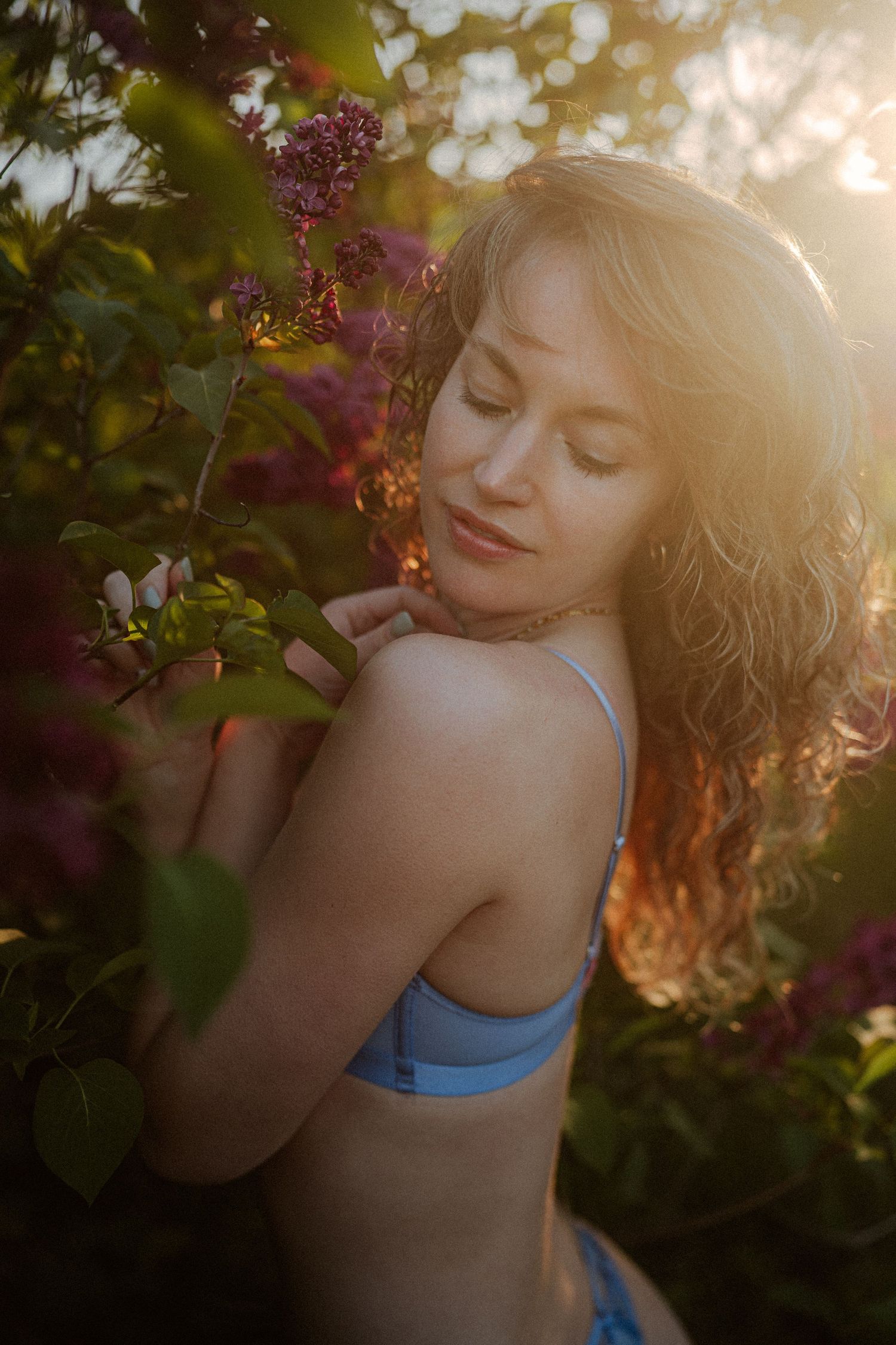 Woman in blue bralette surrounded by blooming lilac bushes in golden sunset light.