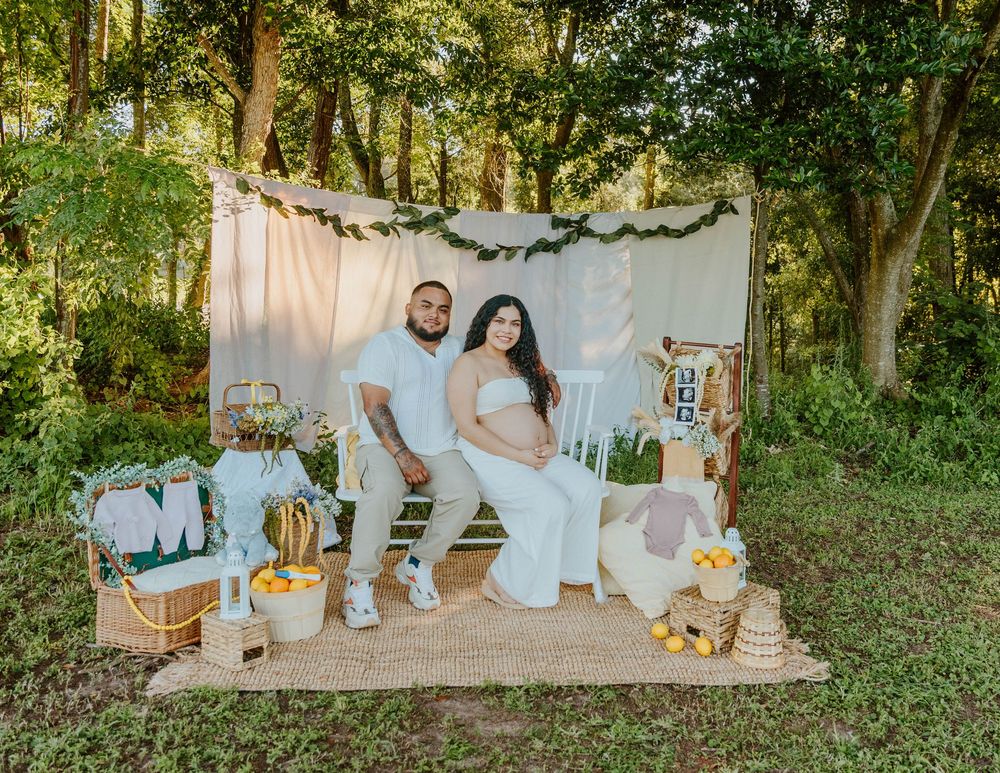 Expecting couple posing on white blanket with decorative props in woodland setting.