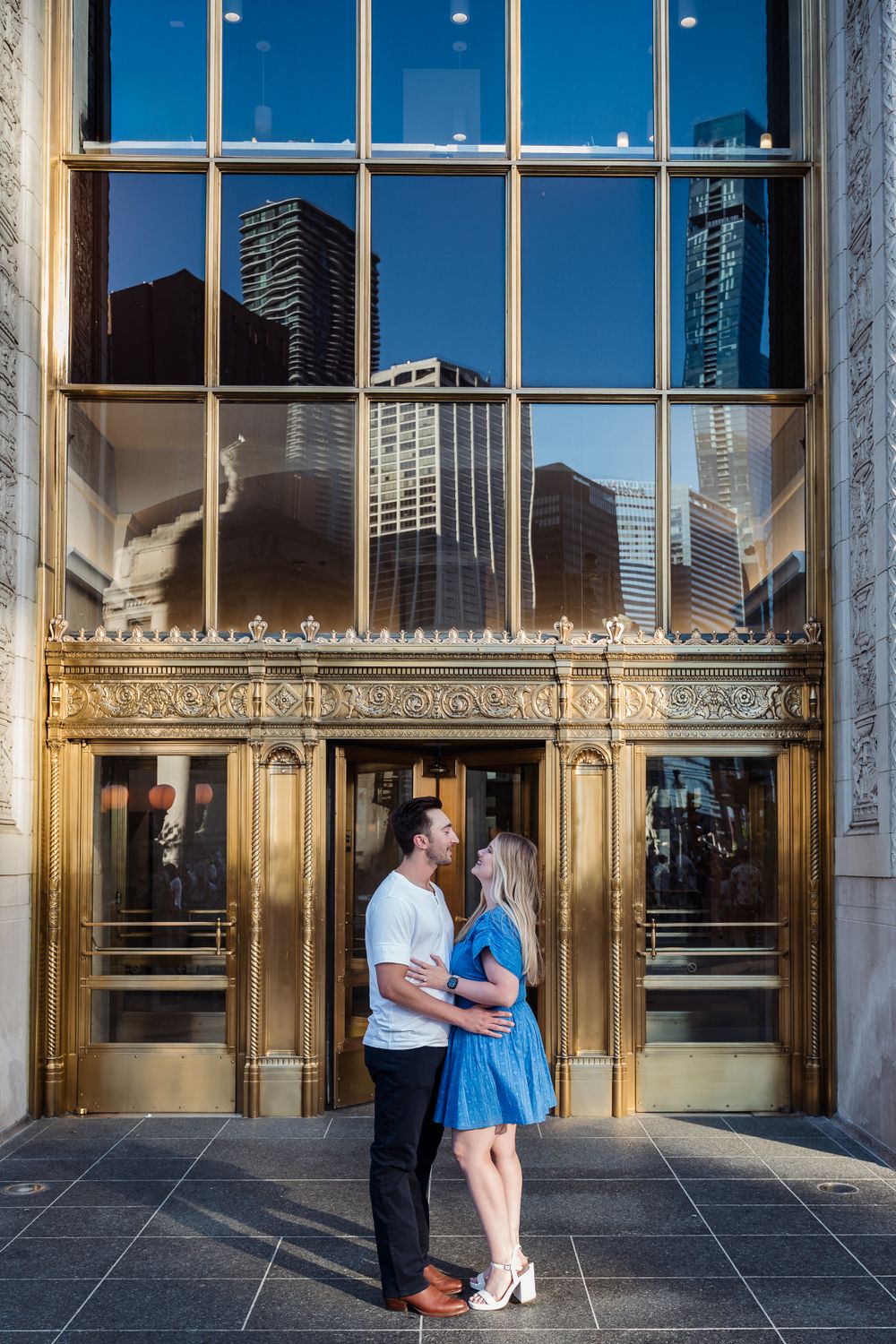 Couple embracing in front of ornate gold-trimmed entrance with city skyline reflection.