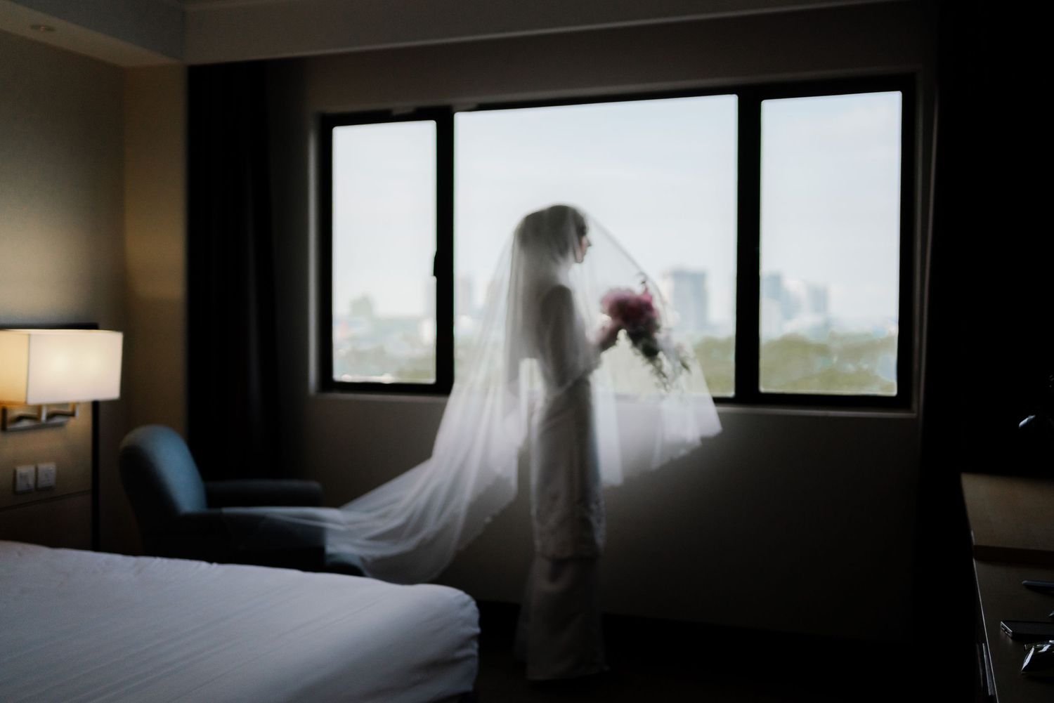 A silhouette of a bride by a window holding flowers in a hotel room.